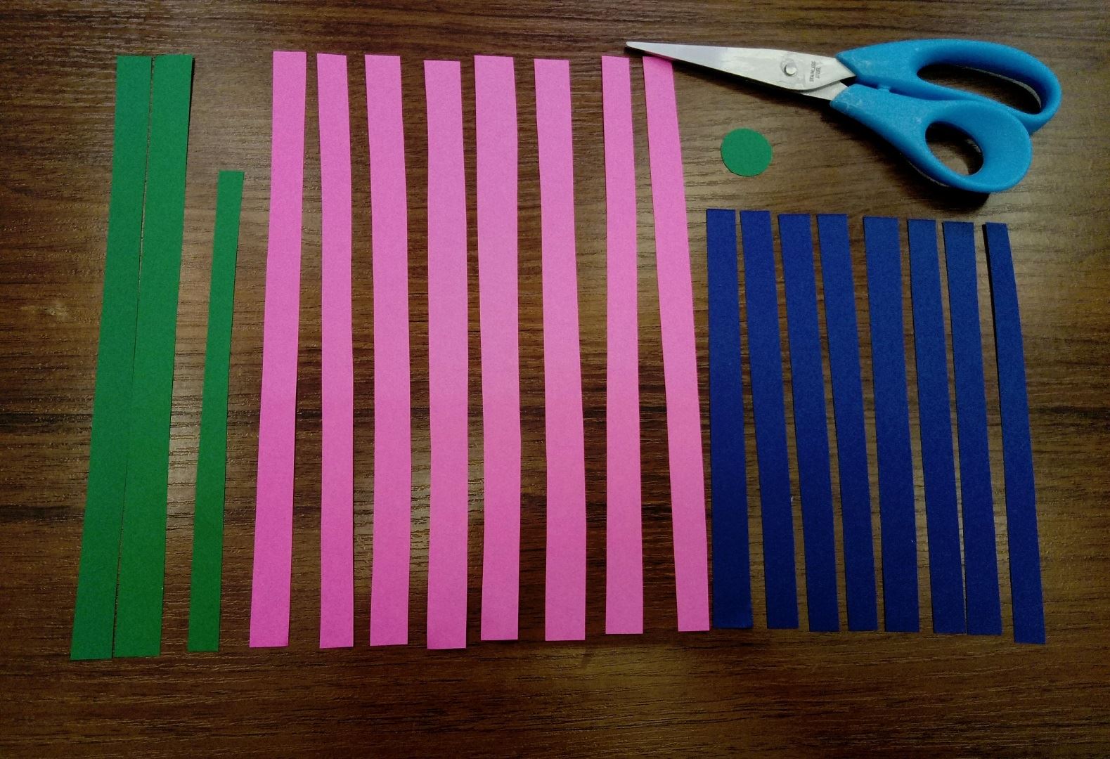 Let's begin from a very simple task, doing a lot of strips. Cut pink paper along into 8 identical strips. Cut horizontally 8 stripes out of the blue paper. Now take the green paper and cut out 2 long strips and one a bit shorter and a small circle. When everything is done, we can continue!