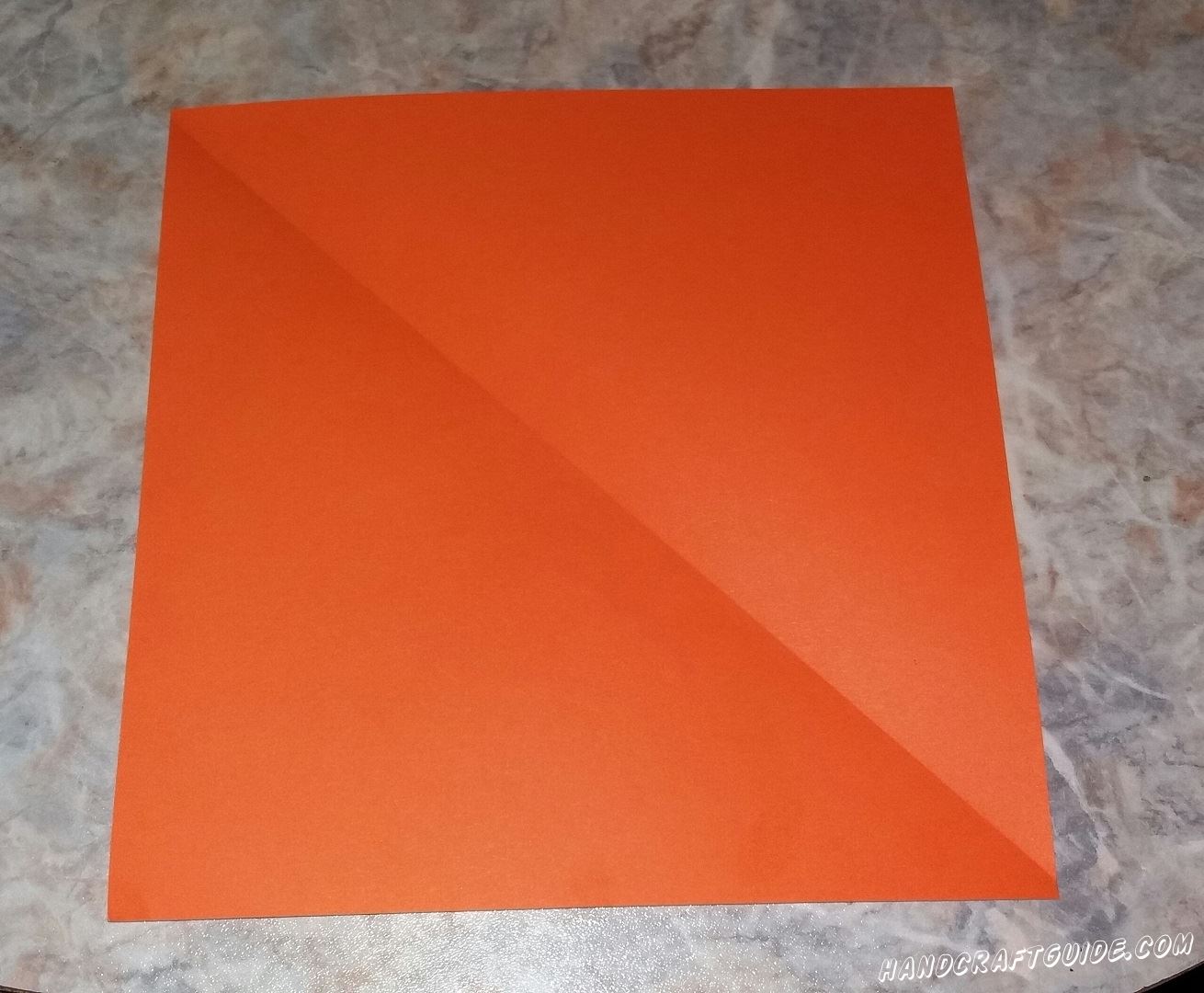 Для начала мы берем оранжевую бумагу и вырезаем ровный квадрат. Затем складываем квадрат пополам в треугольник.