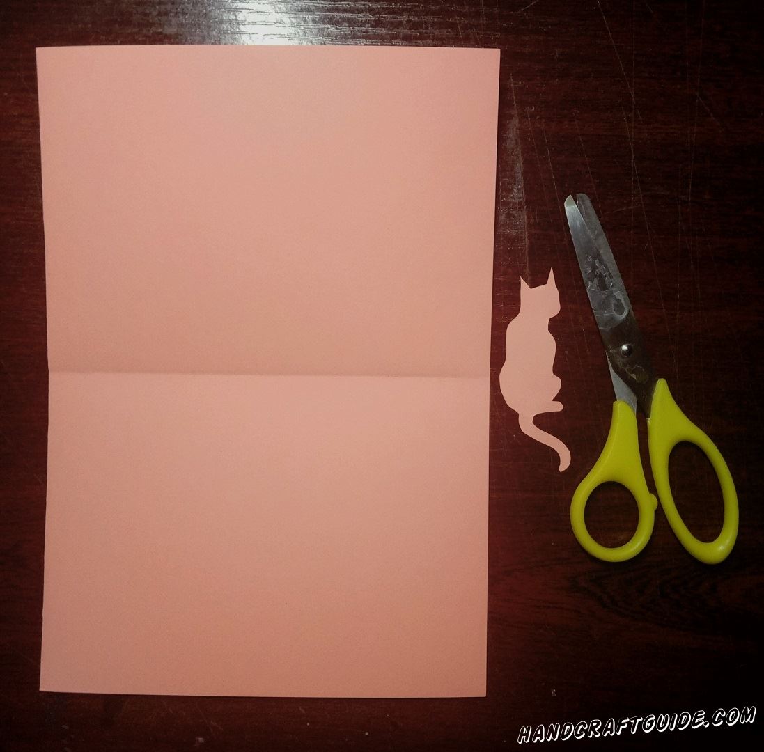 Берем два листа одинакового цвета. На одном из них мы рисуем образ кошечки и вырезаем его. Второй лист мы сгибаем пополам