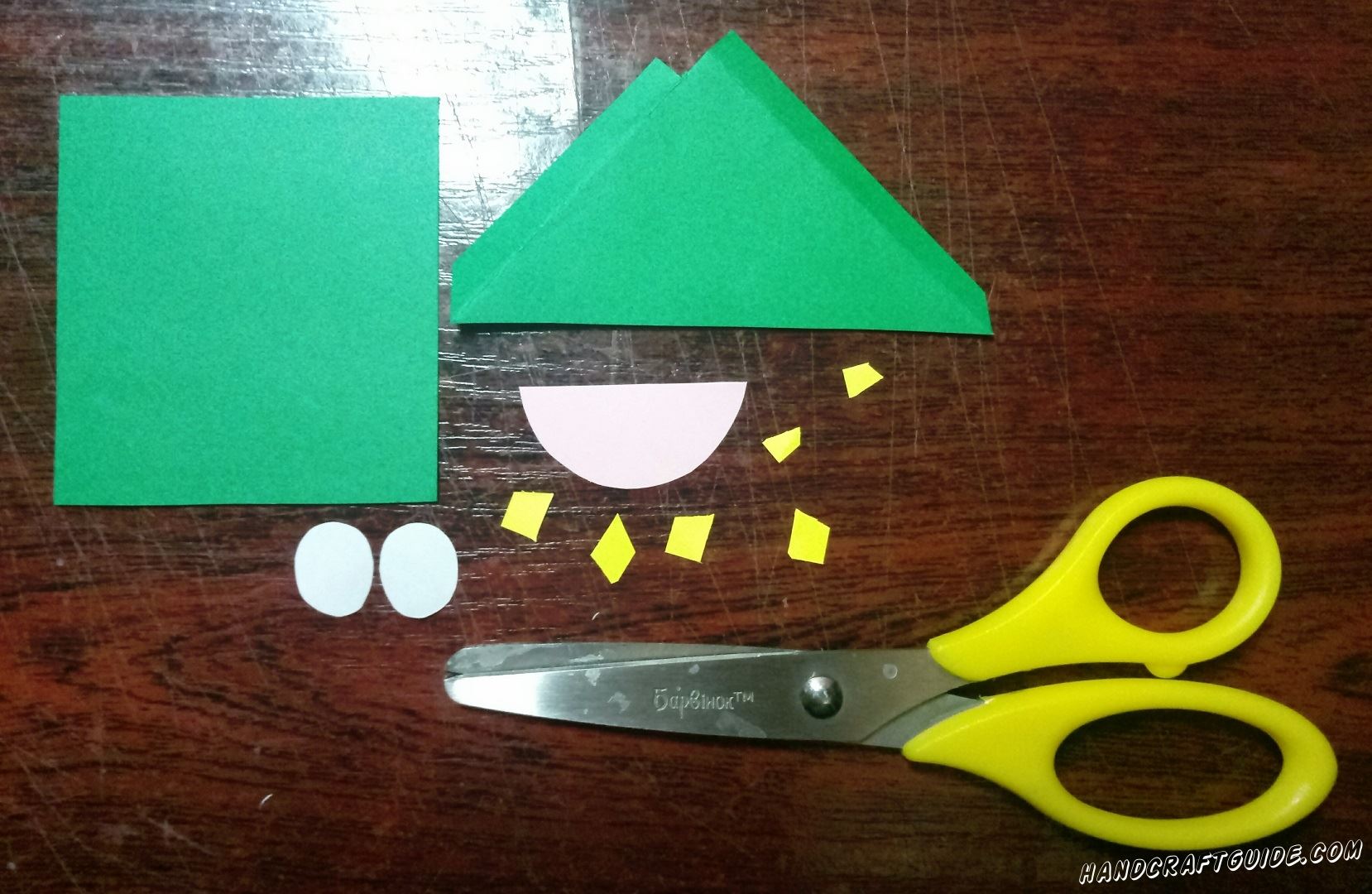 Вырезаем зелёный бумажный треугольник и квадрат. Также нам понадобится светло-розовый ротик, два белых глазика и украшения из маленьких жёлтых четырёхугольных фигурок, как показано на фото